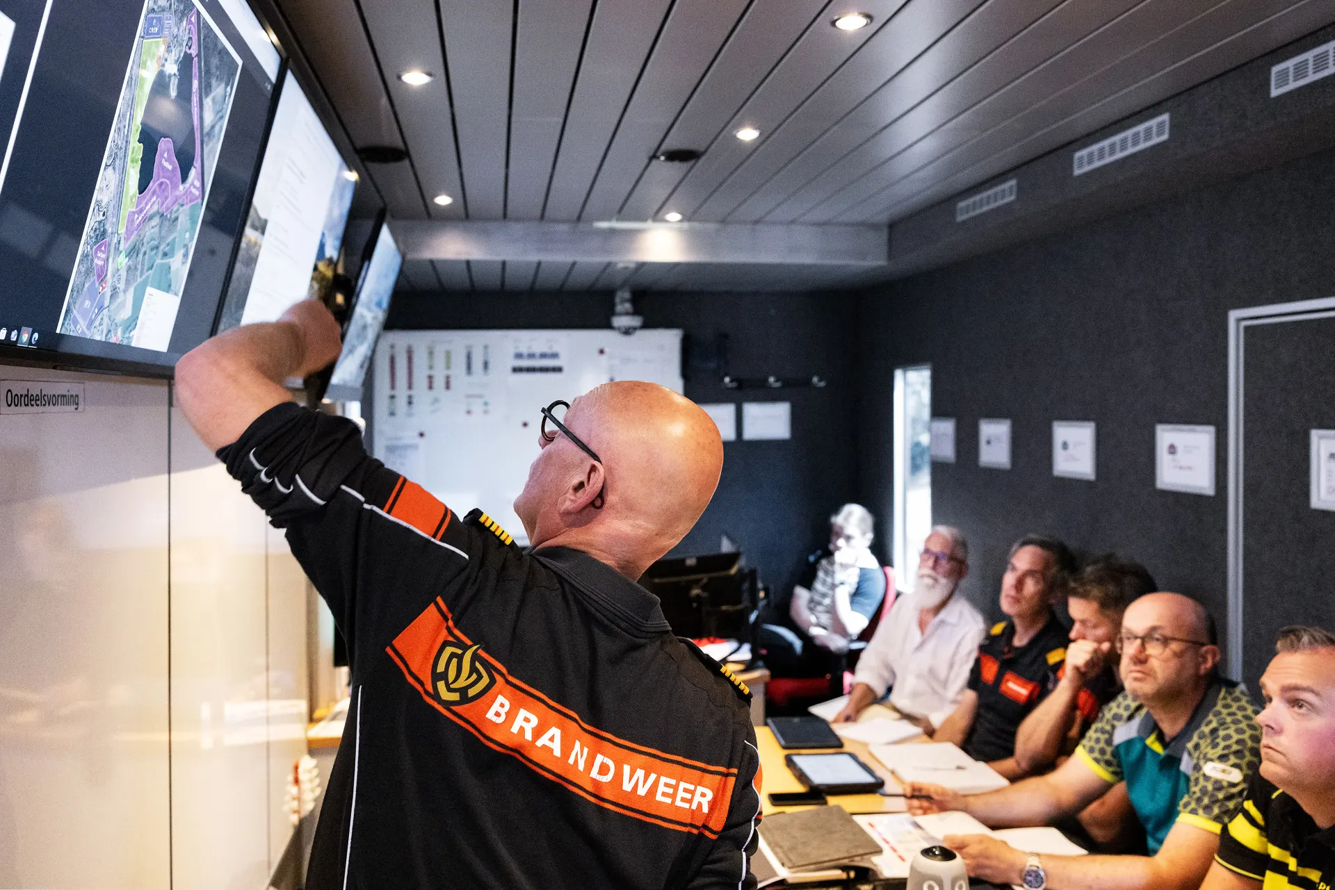 Een commandovoerder wijst op een scherm met een kaart tijdens een oefening in een commandoruimte, met teamleden aan tafel