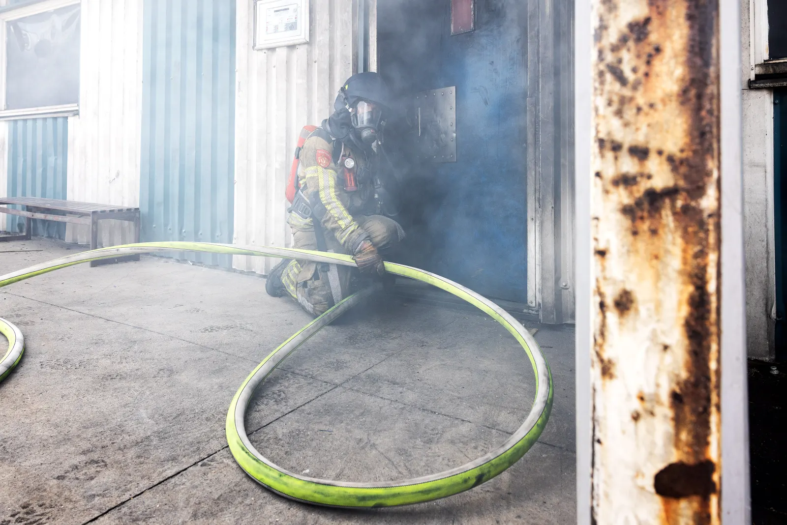 Brandweerman met ademlucht hurkt bij een deur in de rook, met een opgerolde brandslang op de voorgrond